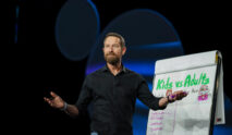 Home 24 A man with a beard, wearing a black shirt and jeans, stands on stage gesturing with open arms next to a flip chart titled Kids vs Adults with drawings and a list written underneath.