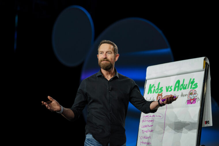 A man with a beard, wearing a black shirt and jeans, stands on stage gesturing with open arms next to a flip chart titled Kids vs Adults with drawings and a list written underneath.