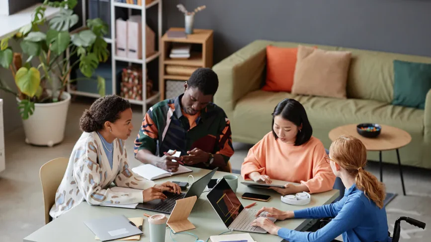 Four people sit around a table working on laptops and tablets in a modern, cozy office with plants and a green sofa in the background. They appear focused and engaged in a network-driven discussion or collaborative project.
