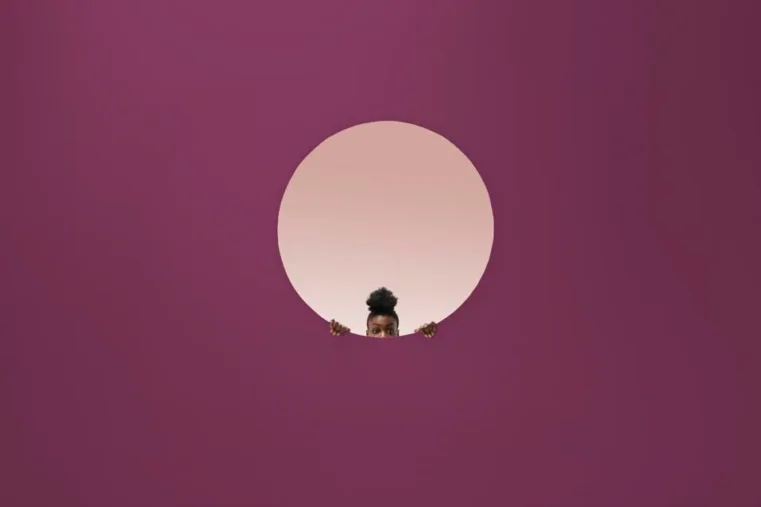 A person peeks through a circular cutout in a large purple wall, showing only their eyes, hands, and hair above the circle’s bottom edge—an image reminiscent of Creativity Speaker Duncan Wardle inspiring curiosity. The background behind the person is light pink.
