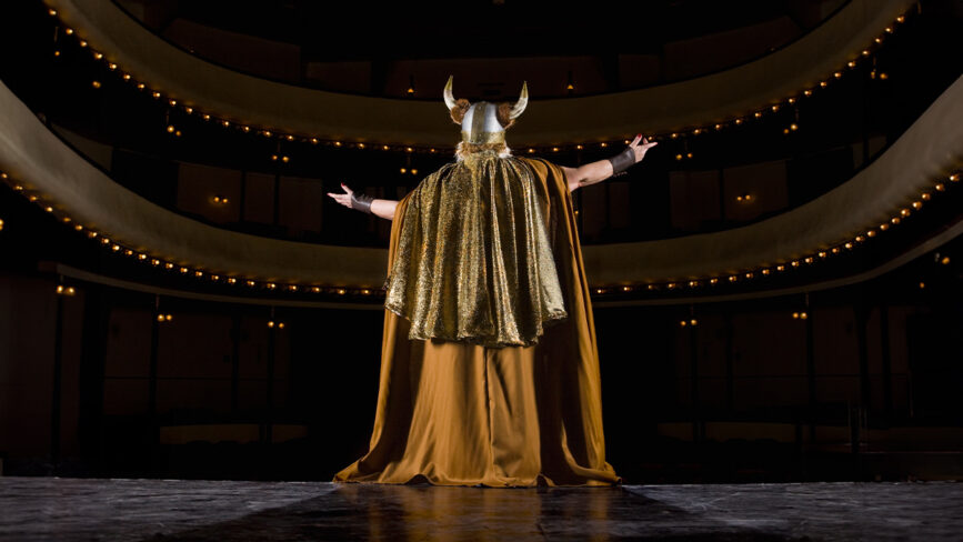A Creativity Speaker in a horned Viking helmet and gold cape stands on a theater stage with arms outstretched, channeling Duncan Wardle’s imaginative spirit before an empty, dimly lit auditorium with glowing lights.