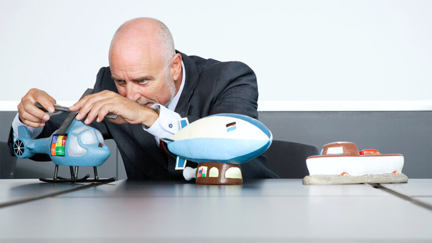Creativity Speaker Duncan Wardle, a bald man in a suit, adjusts a model helicopter on a table, surrounded by model vehicles including a blimp and a boat.