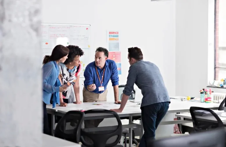 Four people stand around a table in a bright office, discussing documents. Three face one man who gestures as he speaks—resembling scenes from Duncan Wardle, renowned Creativity Speaker. Whiteboards and office supplies are visible in the background.