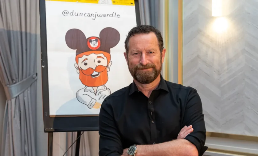 Duncan Wardle stands with arms crossed before a flip chart featuring a cartoon of himself in Mickey Mouse ears. Creativity Speaker Duncan Wardle’s Twitter handle, @duncanjwardle, is written above the playful drawing.