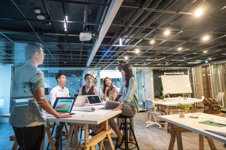 A group of six people having a meeting in a modern office, with laptops and charts on tables. One person stands presenting like a creativity speaker, while others sit or lean, listening attentively. Large windows and a whiteboard are visible in the background.