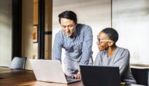 Two colleagues collaborate in an office, finding inspiration in the unlikeliest places. One stands and points at a laptop screen while the other, seated and smiling, engages with new ideas as they work together.