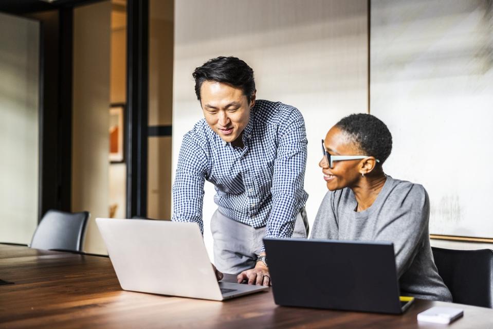 Two colleagues collaborate in an office, finding inspiration in the unlikeliest places. One stands and points at a laptop screen while the other, seated and smiling, engages with new ideas as they work together.